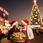 Joyful couple proudly holding a shrimp basket with hats and aprons beside a seashell Christmas tree in a sunset market.