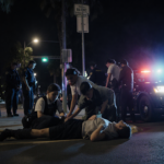 Track student kneeling to tend injured teammate with flashing police lights above the damaged car