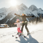 Mother and daughter skiing down a snowy slope at Big Bear Mountain Resort with warm sunlight and snow‑covered peaks.