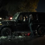 Black G-Wagon rammed into getaway car with shattered glass and neon lights reflecting on wet pavement.