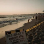 Beachgoers sunbathing with golden light on skin and warning signs near dark waterline.