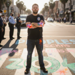 Jonathan Hale stands with arms crossed on the crosswalk painting at Kelton with Vision Zero logo on pavement.