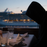Seated figure in dimly lit cruise ship table with overturned drinks and scattered napkins while a blurred ship appears throug