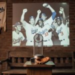 Baseball glove sits open on pedestal with World Series trophy beside it and Dodgers celebration projected onto wall.