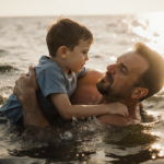 Father holding 7‑year‑old son above water after kayak capsizes with warm sun glow
