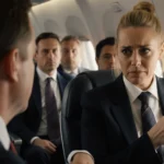 Amy Poehler sits across from a stern first‑class passenger with a coffee cup and window view of blurred cabin.