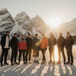 Athletes standing together on a frozen lake with snow‑covered Alps and golden sunset light.