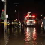 Resident walking toward rescuers with emergency responders and sandbag near Christmas tree on pole in flooded street