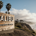 Wooden sign announcing Malibu Unified School District stands on a sunny Malibu shoreline with palm trees and bright blue sky