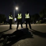 Young person lying on ground with police officers in reflective vests and nighttime cityscape glow