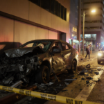 Mangled car lies against modern building with yellow police tape and emergency lights illuminating the nighttime scene.