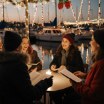 People singing carols with winter coats and golden light from festive boats reflecting on water
