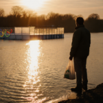 Person standing at flood edge holding reusable bag looking over water with golden sunset and blurred Olympic rink in backgrou