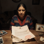 Widow sits with open government letter showing stamp and worn photographs in warm light