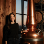 Jobeth Devera stands beside a distillery still with her hand on the wooden wall, light bathing whiskey barrels in background.