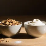 Two bowls of granola and yogurt resting on a wooden table with scattered sugar packets in soft warm light