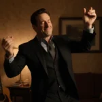 Jon Hamm dancing with eyes closed and one hand raised while holding a wine glass in a warm dim living room.