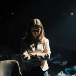 Kristin Cabot stands with crossed arms and downward gaze on dimly lit stage with empty seats and scattered Coldplay merch