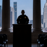 LAPD officer standing at podium with empty City Hall chamber and city lights outside
