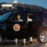 Two men lean out of a black SUV with a stylized L and basketball logo on a dark LA street at dusk near Hollywood Hills
