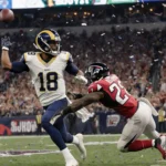 Rams quarterback throwing a pass with defender tackling and receiver sprinting down sideline in stadium crowd with confetti.