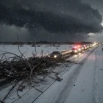 Stranded cars glowing with warm headlights on a snow-covered highway with tangled downed power lines and looming storm cloud.