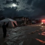 Lone figure gazing at devastation with muddy water swirling around abandoned homes and a rescue vehicle with sirens