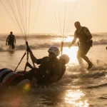 Two paragliders partially submerged are rescued by deputies with sunset light bathing the scene and a golden sky.