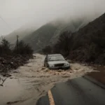 Crushed car lying submerged in muddy water with scattered trees and misty mountains in background