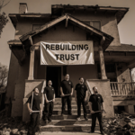 Five men in black tools look up at partially rebuilt Altadena house with banner saying REBUILDING TRUST near Eaton debris.