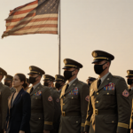 Uniformed soldiers marching with a civilian figure beside them and a faded American flag fluttering in background