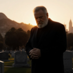 Rob Reiner stands at a gravesite with a small marker and the Los Angeles skyline in a warm sunset glow while his eyes look do