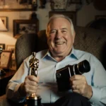 Rob Reiner sits holding a film camera and an Oscar with warm lamp light and nostalgic film memorabilia.