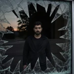 Claudio Manuel Neves Valente reflected in shattered windowpane with scattered glass and silhouetted New Hampshire trees
