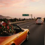 Packed car on Southern California highway with holiday decorations drives past bright orange convertible near sunset sky.