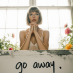 Taylor Swift sits and clasps her hands with light pouring through a large window beside lush greenery and a faded chalkboard