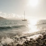 Solitary sailboat glides across calm Channel Islands water with gentle wave against Ventura County rocks under warm sun