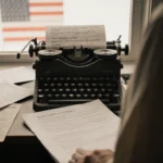 Person standing before typewriter with job listings and resumes in background and open contract on desk with flag at window