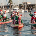 Participants paddleboarding through calm waters of Ventura Harbor Village with colorful elves and Santa hats in background