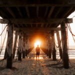 Beachgoer standing on pier at dusk with warm orange glow and weathered wooden slats and tangled cables