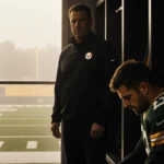Aaron Rodgers sits on locker room bench with head bowed while Coach Tomlin stands before him and football field shows through