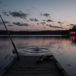 Abandoned fishing rod lies on wooden dock at dusk with rippling lake water and faint bowling alley visible on shore