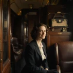 Lady Caterham sits alone in train compartment with vintage luggage and warm golden lighting