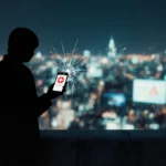 Person stands at city edge with cracked phone showing emergency app and shadowed face looking over glowing skyline