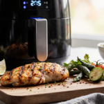 Air‑fried chicken breast being plated on a wooden cutting board with fresh ingredients around it and an air fryer glowing in
