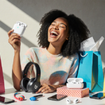 Woman holding up new AirPods with smiling eyes closed amid colorful Beats earbuds and Apple devices in bright natural light