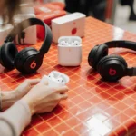 Shoppers examine AirPods and Beats headphones on colorful tablecloth with deals signs showing savings