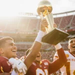 Alberto Mendoza holds the Heisman Trophy aloft with Max cheering near Indiana University stadium under sunlight