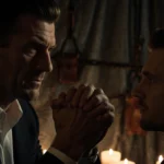 Two men hold hands with fingers intertwined and candlelight revealing leather restraints and BDSM equipment in the background