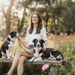 Amanda Batula sits on a bench holding a leash to a dog while stroking a dog beside her with sun filtering through green park